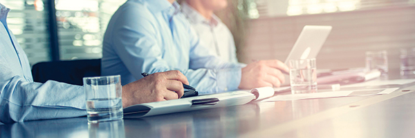 Man sitting at a desk taking notes with another man working on a laptop sitting next to him.